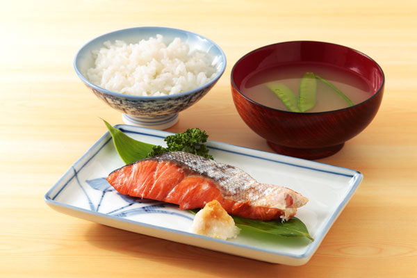 パセリと大根おろしを添えて盛りつけた紅鮭の皿の背後にご飯と味噌汁を置いた画像。背景は明るい木目のテーブル