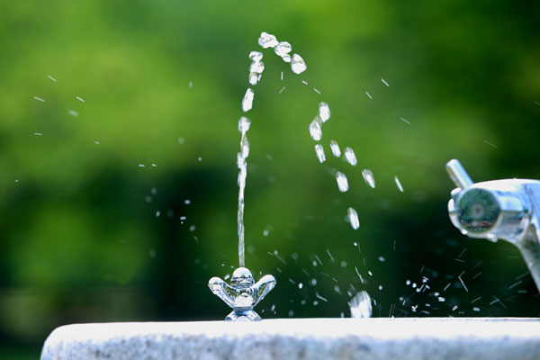 アウトフォーカスした木立の緑を背景にして涼やかに水玉になって吹き上水飲み場も水。